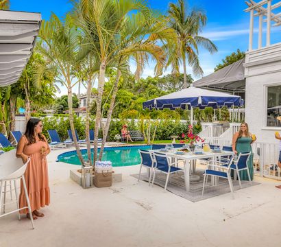 Sunny tropical backyard pool patio with palm trees, navy umbrella, outdoor dining set, lounge chairs and guests enjoying drinks.