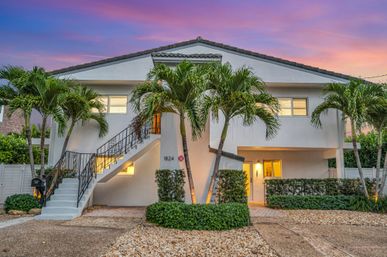 Modern two-story coastal home under a pink-purple sunset, exterior staircase leading to an upper level, swaying palm trees, warm lit entryway, gravel driveway and manicured hedges.
