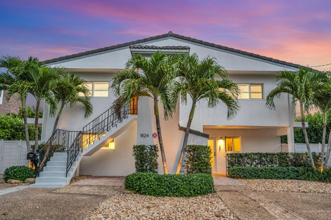 Modern two-story coastal home under a pink-purple sunset, exterior staircase leading to an upper level, swaying palm trees, warm lit entryway, gravel driveway and manicured hedges.