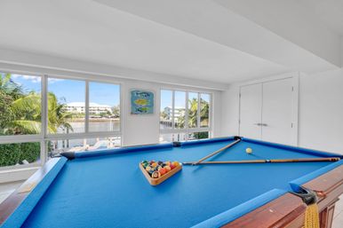 Sunny waterfront game room with bright blue-felt pool table, racked billiard balls and cues, floor-to-ceiling windows framing palm trees and a canal-front coastal neighborhood.