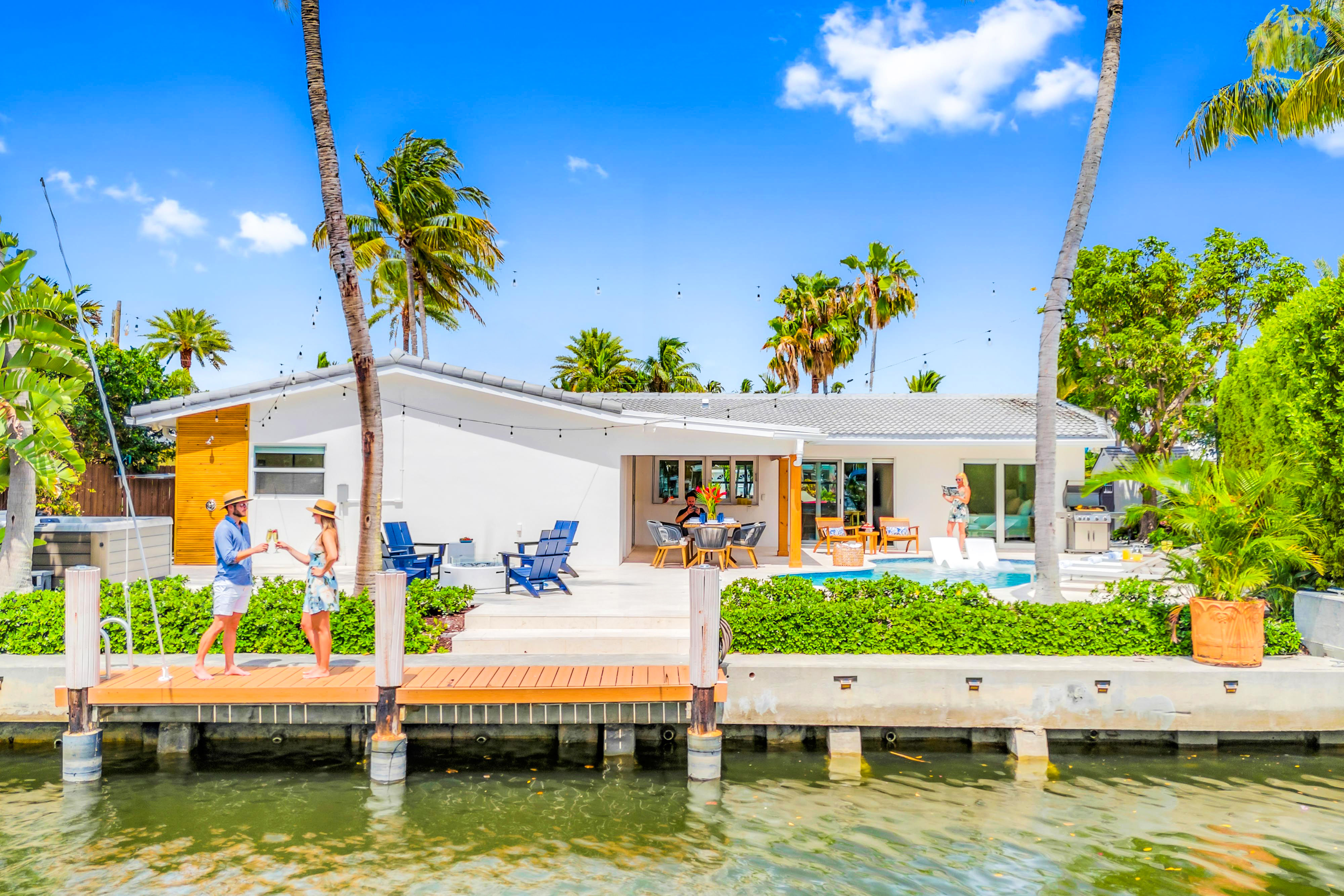 Sunny waterfront modern home with pool and patio, palm trees and wooden dock on a tropical canal, two people toasting near the water