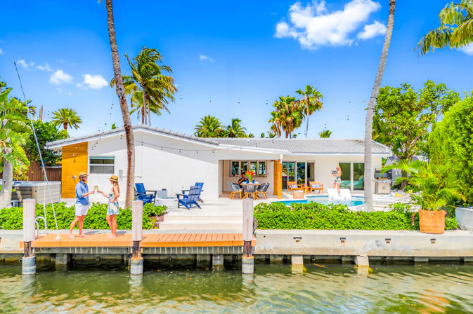 Sunny waterfront modern home with pool and patio, palm trees and wooden dock on a tropical canal, two people toasting near the water