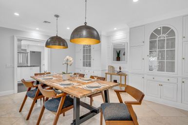 Modern dining room with wooden farmhouse table set for six, mid-century wooden chairs with blue cushions, oversized hammered metal pendant lights, white built-in cabinetry and polished stone floor.
