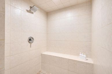 Neutral beige tiled walk-in bathroom shower with built-in bench, square rainfall showerhead, chrome control handle, and three white pump bottles for a spa-like modern look.