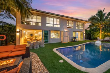 Sunset-lit two-story tropical waterfront home with glowing interiors, backyard pool, palm trees, outdoor dining, cozy lounge seating and fire pit on a manicured lawn.