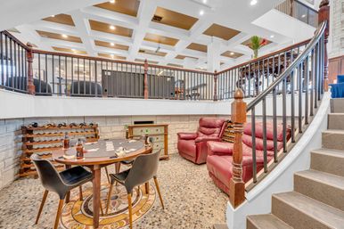 Cozy sunken home basement recreation room with round poker table and chairs, wall wine rack, red leather recliners, pebble stone floor, wooden staircase and iron railing beneath a coffered ceiling.