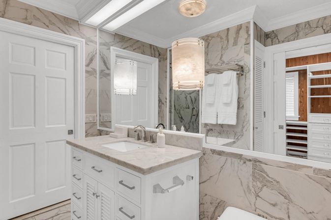 Spa-like bright marble bathroom with white vanity and marble countertop, undermount sink, chrome faucet, large mirror, cylindrical pendant light, towel rack with folded towels, and doorway to a wooden walk-in closet.