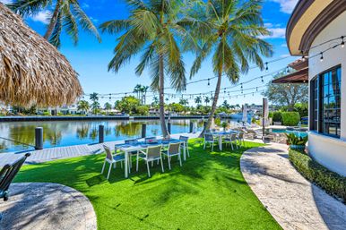 Canal-front tropical backyard patio with palm trees, string lights, outdoor dining tables on green lawn, poolside lounge area and boat dock under a bright blue sky.