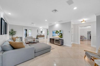 Bright open-concept modern living room with light-gray sectional chaise, beige tile floors, TV on a media console, round wall mirror, armchair and French doors in a suburban home.