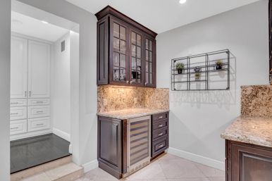 Stylish home wet bar with dark wood glass-front cabinets, under-cabinet lighting, granite countertops and backsplash, built-in wine fridge and a wall-mounted metal shelf with small potted plants; white pantry visible through doorway.