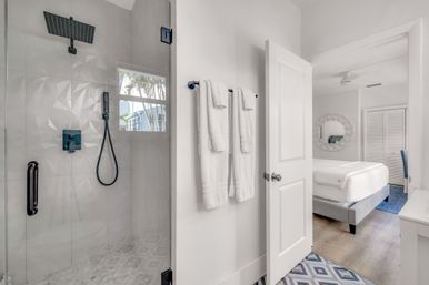 Bright modern coastal-style bathroom with glass walk-in shower, matte black rain and handheld shower fixtures, white towels on wall hooks, geometric tile floor, open door leading to airy bedroom with white bed and round mirror and palm trees visible through shower window.