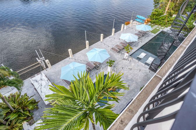 Overhead balcony view of a waterfront pool deck with light-blue umbrellas, lounge chairs, string lights, palm trees, and kayaks