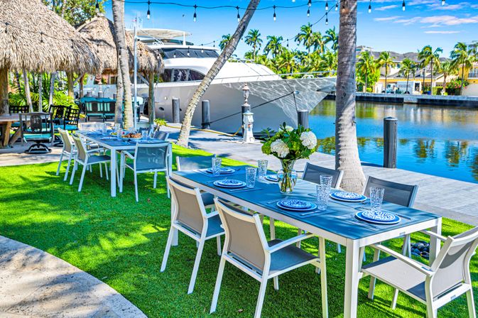 Tropical waterfront dining scene with blue-topped tables set with blue-and-white plates, glassware and a white hydrangea vase on a green lawn beside a marina featuring a luxury yacht, palm trees, thatched tiki huts and string lights.