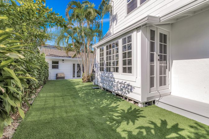 Sunny residential backyard with white house and sunroom, glass French doors, artificial turf lawn, tall palm trees and a dense hedge.