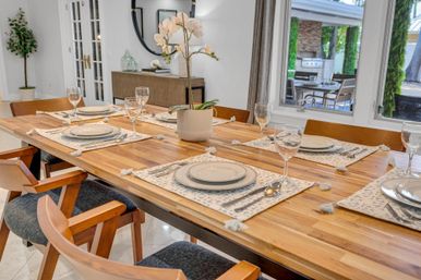Sunlit modern dining room with a long wooden table set for six — patterned placemats, white dinnerware, wine glasses and a potted white orchid centerpiece, large windows overlooking a patio.