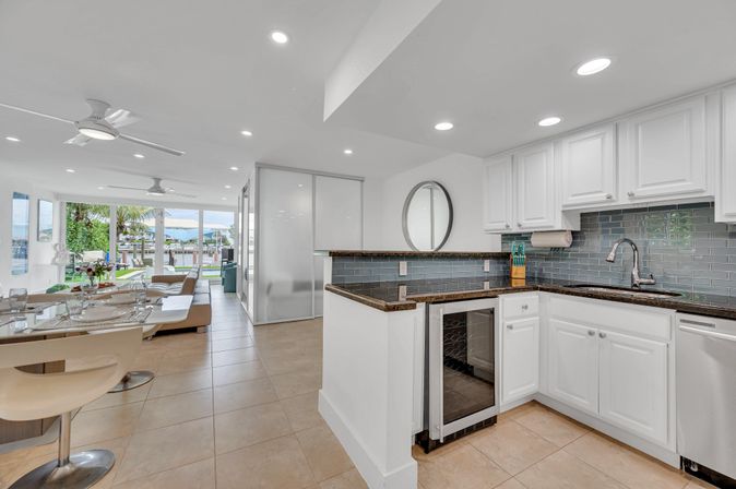Sunny waterfront condo with open-plan kitchen and dining area, white cabinets, dark granite countertops, blue glass subway-tile backsplash, breakfast bar with stools, tile floors, and floor-to-ceiling windows revealing a marina and palm trees.