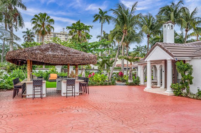 Resort-style tropical patio with a thatched tiki bar and bar stools, red stamped concrete, white columned pool house, lush palm trees and vibrant landscaping under a blue sky.