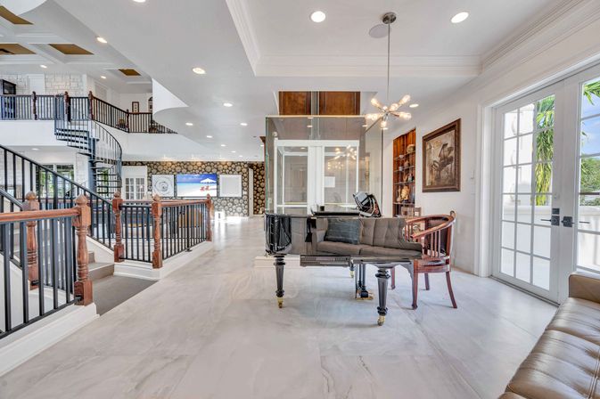 Bright tropical luxury foyer with glossy black grand piano, curved wrought-iron spiral staircase, marble floors, mid-century chandelier and French doors opening to palm trees.
