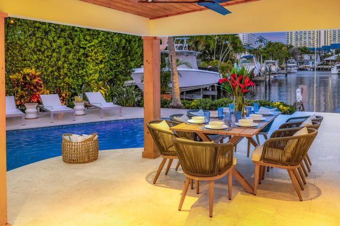 Waterfront tropical patio by a canal with pool, docked boats, and a poolside dining table set for eight with wicker chairs and red tropical flowers.
