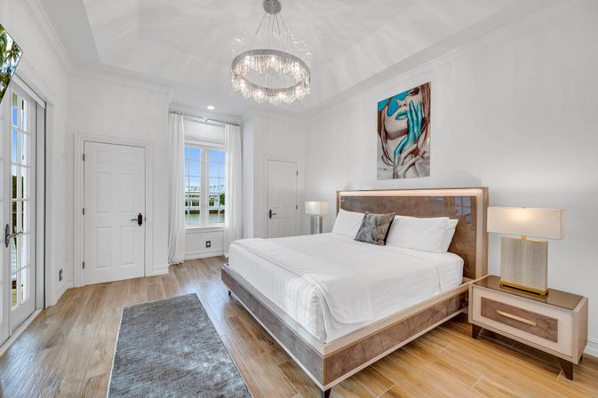 Bright waterfront master bedroom with king bed, sparkling crystal chandelier, wood-look floors, modern nightstands and contemporary artwork — airy coastal luxury.