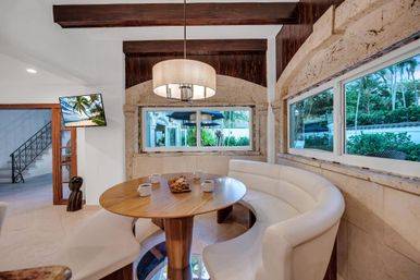 Cozy tropical breakfast nook with curved white banquette around a round wooden table, pendant chandelier, stone arched walls and windows overlooking lush garden