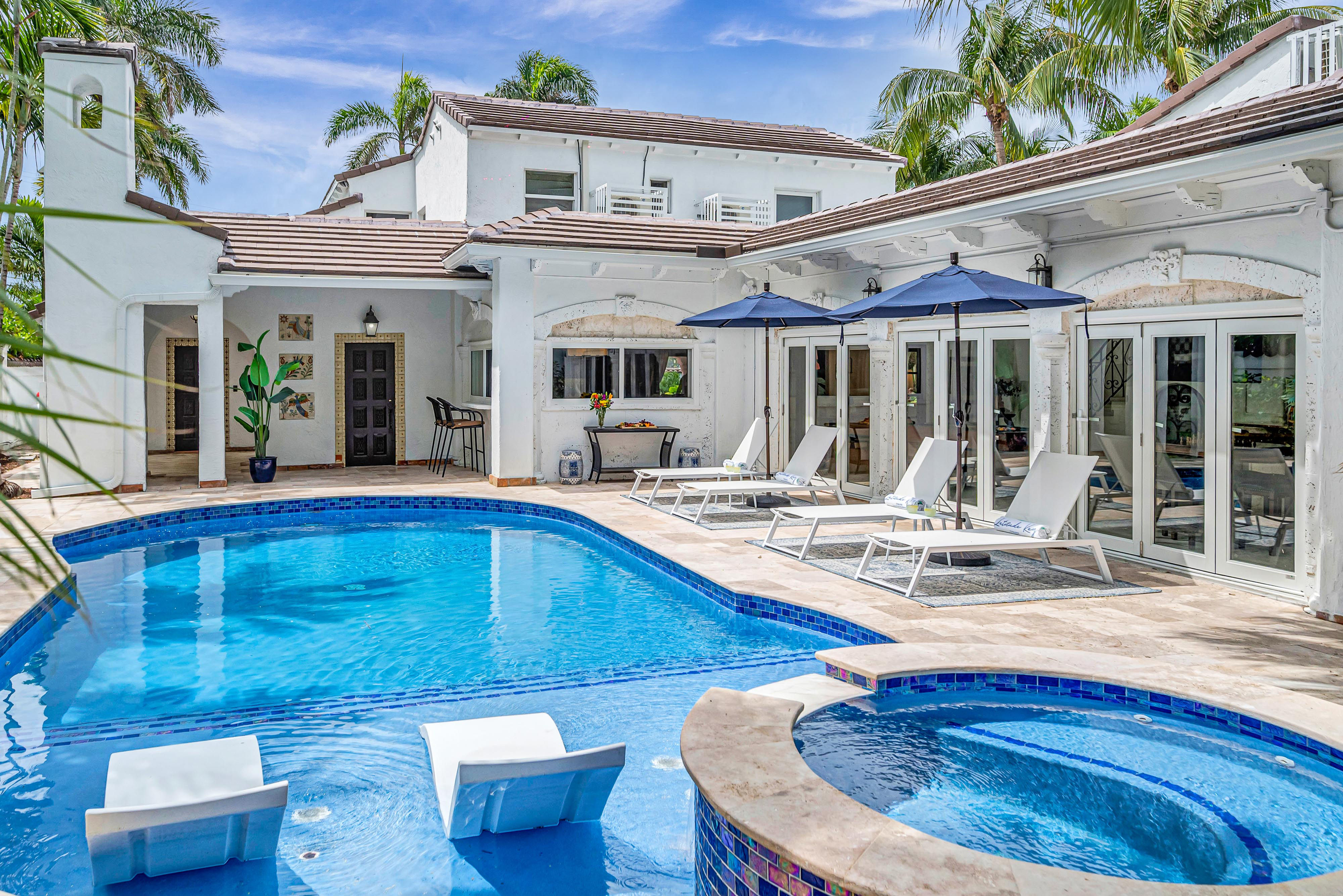 Tropical luxury backyard with blue-tiled swimming pool and circular spa, white Mediterranean-style villa patio lined with sun loungers, navy umbrellas, palm trees, and poolside seating.