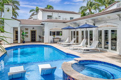 Tropical luxury backyard with blue-tiled swimming pool and circular spa, white Mediterranean-style villa patio lined with sun loungers, navy umbrellas, palm trees, and poolside seating.