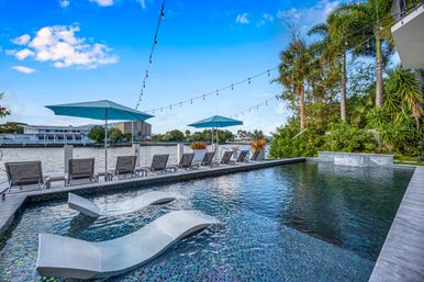 Sun-ready waterfront infinity pool with in-pool lounge chairs, row of sunbeds and blue umbrellas along a canal, palm trees and string lights overhead