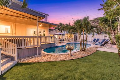 Sunset backyard with kidney-shaped swimming pool, tropical palm trees, pebble border and reclining loungers beside a covered wooden deck and glowing patio lights