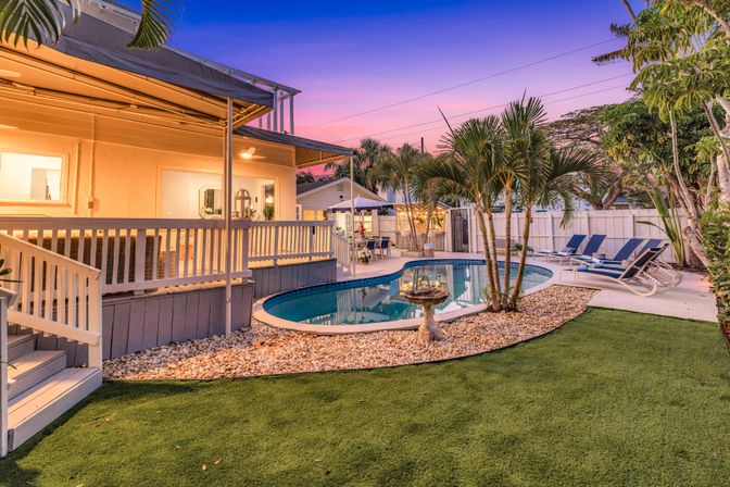 Sunset backyard with kidney-shaped swimming pool, tropical palm trees, pebble border and reclining loungers beside a covered wooden deck and glowing patio lights