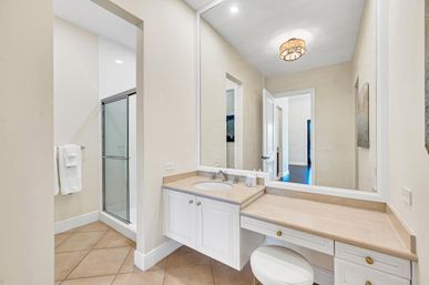 Bright residential bathroom with large wall mirror, single-sink white vanity and makeup counter with cushioned stool, glass-enclosed shower, beige tile floor and neutral walls
