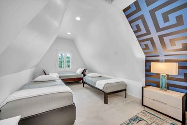 Bright modern attic bedroom with sloped white ceilings, two twin beds and a daybed, bold blue geometric accent wall, nightstand with lamp, and shuttered window