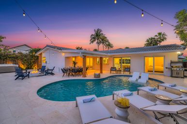 Sunset backyard oasis with palm trees and kidney-shaped pool, tiled patio with lounge chairs and in-pool loungers, string lights, fire pit, outdoor dining area and grill.