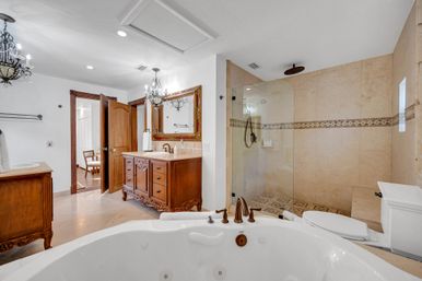 Resort-style luxury master bathroom with a jetted soaking tub in the foreground, ornate wooden vanity and framed mirror, glass-enclosed walk-in shower with rainfall head and decorative tile border, chandeliers and marble tile floors.