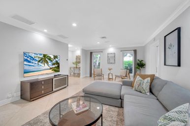 Bright modern coastal-inspired living room with light gray sectional sofa, glass coffee table, wall-mounted TV displaying a tropical beach, marble tile floors, two accent chairs by sliding glass doors, plants and minimalist wall art