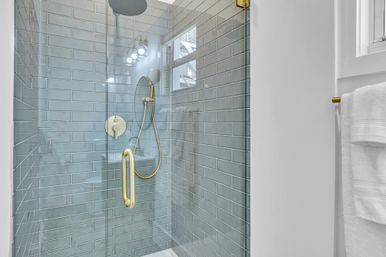 Sleek glass-enclosed shower with blue-gray subway tile, brass rain and handheld shower fixtures, gold handle and white towel on nearby wall
