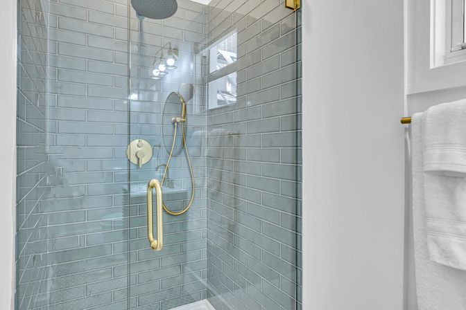 Sleek glass-enclosed shower with blue-gray subway tile, brass rain and handheld shower fixtures, gold handle and white towel on nearby wall
