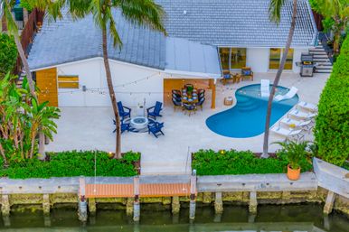 Aerial view of a waterfront modern backyard with a kidney-shaped turquoise pool, poolside loungers, outdoor dining table, fire pit surrounded by blue chairs, string lights, palm trees and a small private dock.