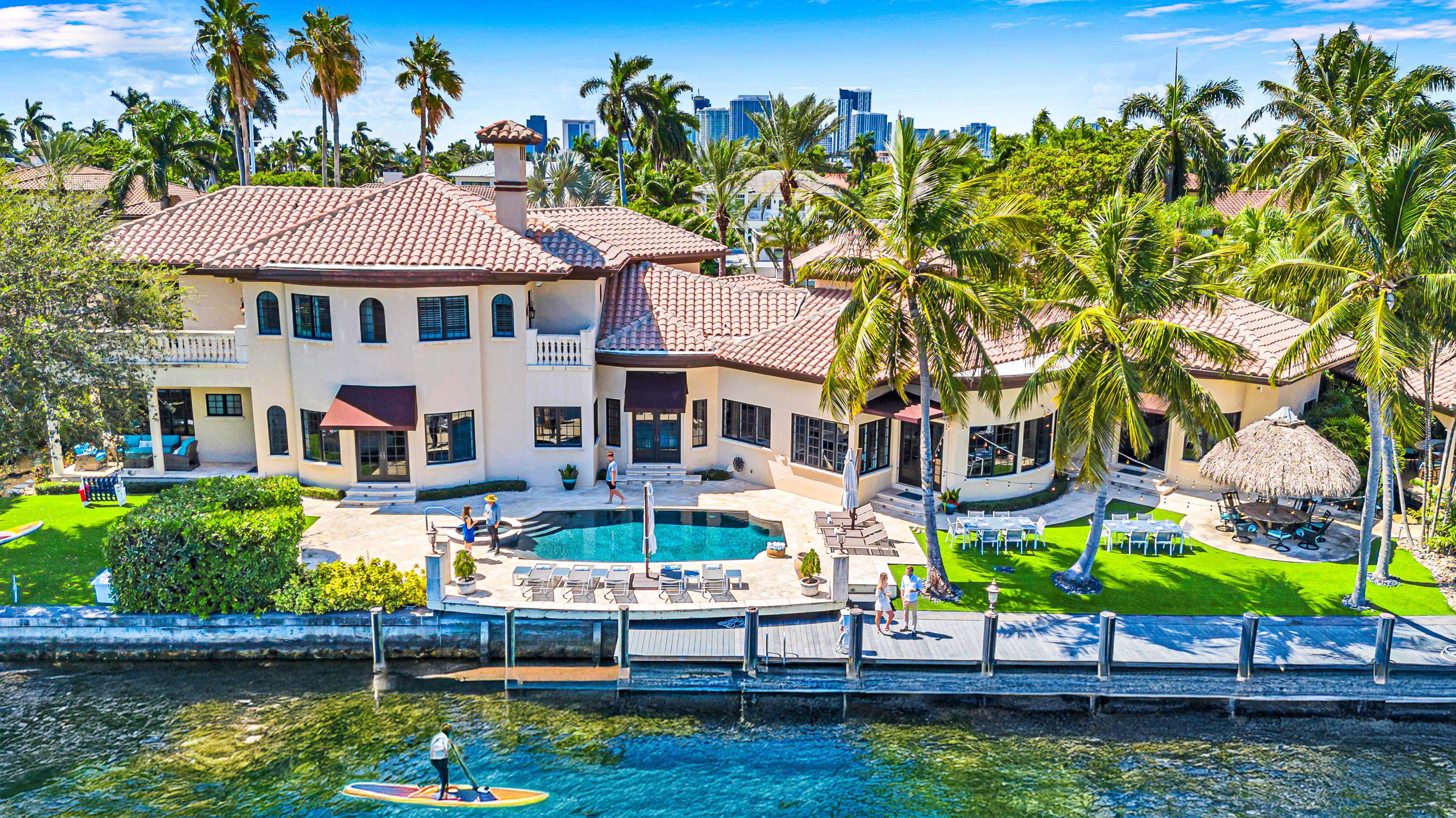 Aerial view of a Mediterranean-style waterfront mansion with pool, palm trees and dock; paddleboarder on blue water and a city skyline in the background.