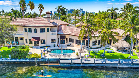 Aerial view of a Mediterranean-style waterfront mansion with pool, palm trees and dock; paddleboarder on blue water and a city skyline in the background.