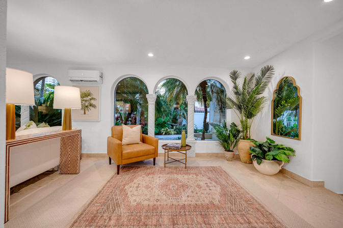 Bright Mediterranean-style living room with three arched windows opening to a tropical garden, tan leather armchair, patterned boho rug, potted palms and gold-framed mirror.