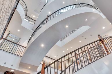 Upward view of a bright modern multi-level atrium with sweeping curved balconies, black metal railings, wooden newel posts and recessed ceiling lights on white walls