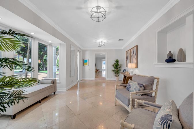 Bright modern foyer and sitting area with marble tile floors, floor-to-ceiling windows revealing palm trees, neutral armchairs and bench, potted plants and minimalist coastal-style decor