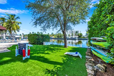 Tropical waterfront lawn with oversized Connect Four and cornhole games, stacked kayaks, palm trees and a canal with boats under a bright blue sky.