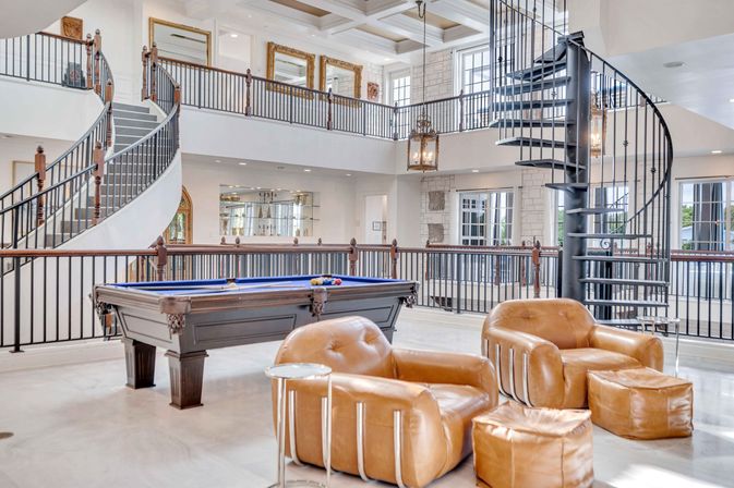 Spacious luxury game room with blue-felt pool table, caramel leather lounge chairs and ottomans, dramatic spiral staircase and curved mezzanine railings in a bright two-story open-plan interior.