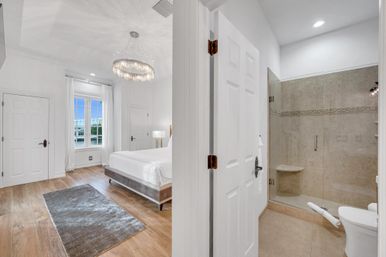 Bright modern coastal bedroom with crystal chandelier, white platform bed, hardwood floors and window with waterfront view; adjoining tiled ensuite bathroom with glass shower and toilet.