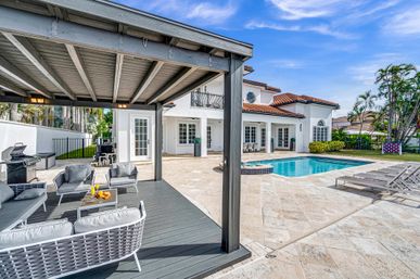 Sunny luxury Mediterranean-style backyard with red-tile roof home, tiled swimming pool and spa, covered gray pergola lounge with cushioned seating, outdoor grill and sun loungers amid tropical palms.