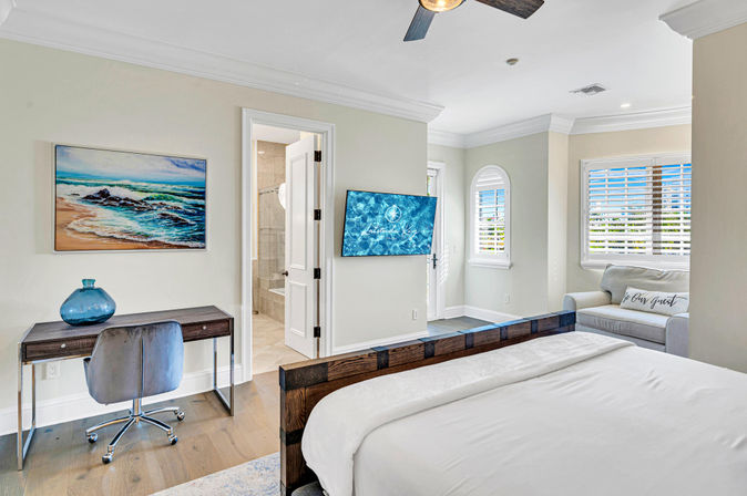 Bright coastal-style bedroom with wooden bed frame and white linens, wall-mounted TV, small desk with blue glass vase under an ocean painting, open ensuite bathroom doorway, and a sunny armchair by shuttered windows.