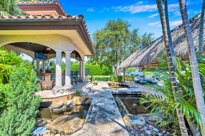 Tropical backyard with stone walkway over a koi-style pond leading to a tiled-roof covered patio with columns and an adjacent thatched tiki hut, palm trees and lounge seating under a bright blue sky.