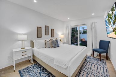 Bright modern coastal bedroom with king-size bed in crisp white linens, blue patterned rugs and accent pillows, bedside lamps and a blue chair, with sliding glass doors opening to a tropical patio with palm trees and outdoor seating.